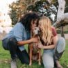 photo of a queer couple, one Black woman and one white woman, each kissing a dog outdoors