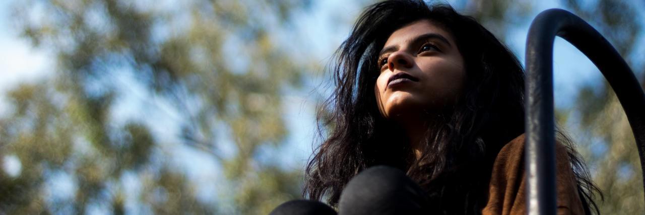 My Experience of Bipolar Disorder Mania, Depression, and Stability photo of a woman sitting with her legs raised to her chest, looking away from camera with blurry background of trees, taken from below