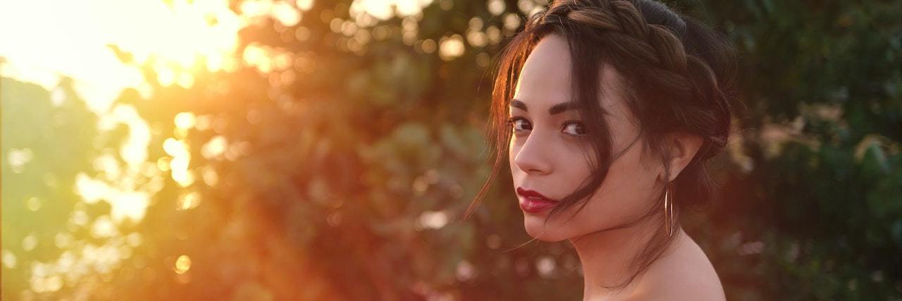What Does It Actually Mean to Have a 'Personality Disorder'? A woman with dark hair pulled back with long bangs framing her face stands next to a bush at sunset