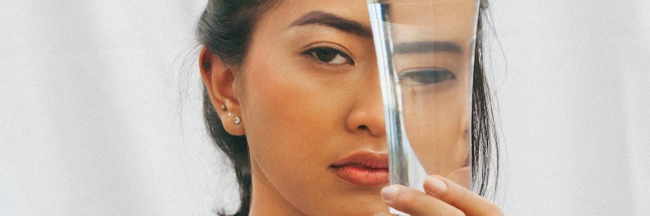 Does PTSD Cause Dissociation? Woman with dark hair wearing a white shirt looks through a glass of water that distorts her features