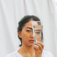 Woman with dark hair wearing a white shirt looks through a glass of water that distorts her features