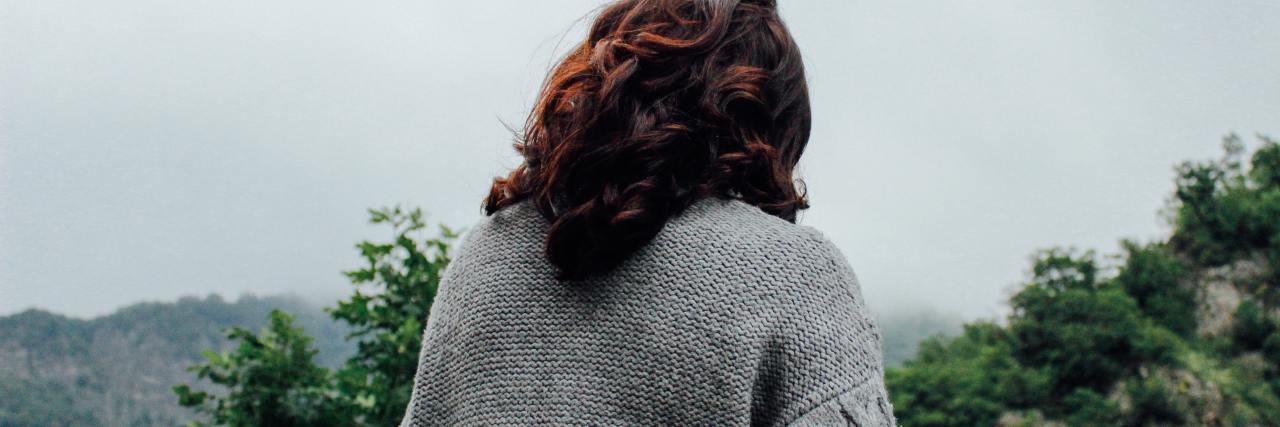 photo of woman in white cardigan taken from behind, looking down with trees in background
