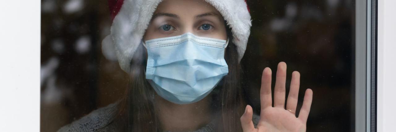 photo of woman behind glass alone wearing Santa hat and coronavirus mask, looking sad