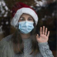 photo of woman behind glass alone wearing Santa hat and coronavirus mask, looking sad