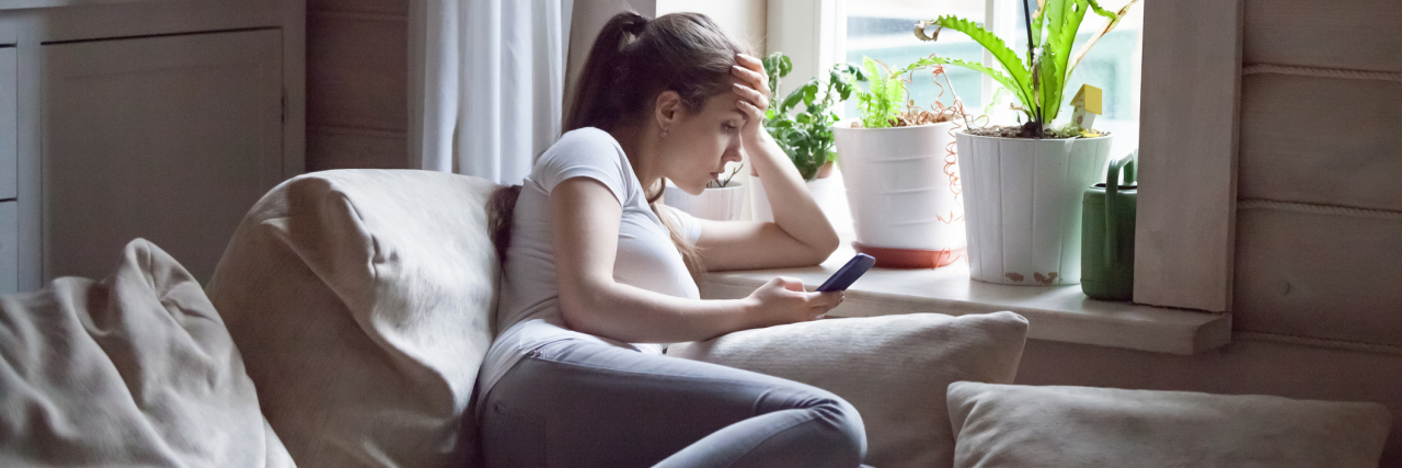 My 'Typical' Day With Borderline Personality Disorder photo of a white woman curled up on the couch upset with one hand on her forehead and the other holding her phone