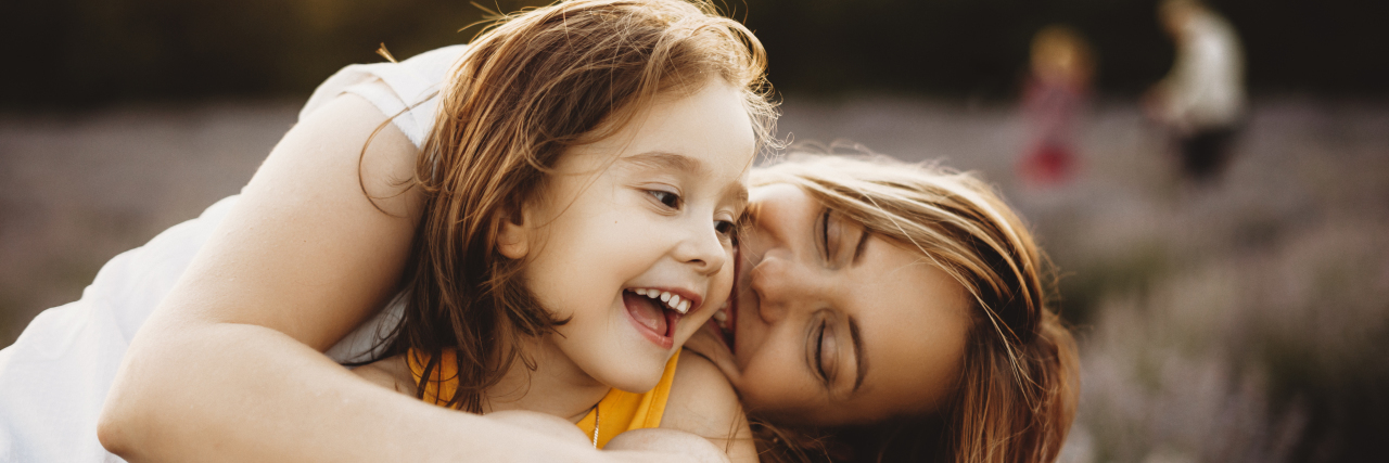Managing Emotions After Your Child's Disability Diagnosis Little girl laughing while being embraced by her mother outdoors in field of flowers.
