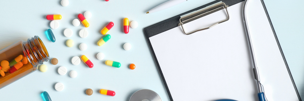 A bottle of meds being slipped next to a stethoscope and a doctor's notepad