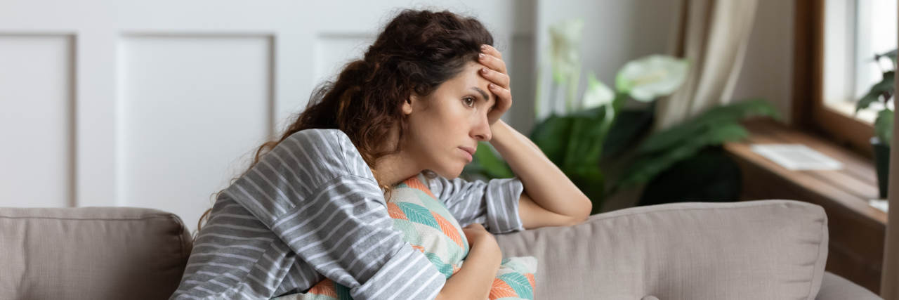 When Migraine Relief Comes, but Adjusting Is Hard A young, white woman with brown hair curled up on the couch with a hand on her head, looking away lost in thought