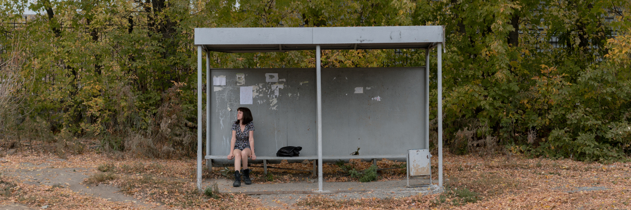I'm Not 'Lucky' That I Can't Drive Because of My Learning Disability Woman in a dress and boots sits at a bus stop.