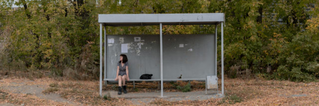 Woman in a dress and boots sits at a bus stop.