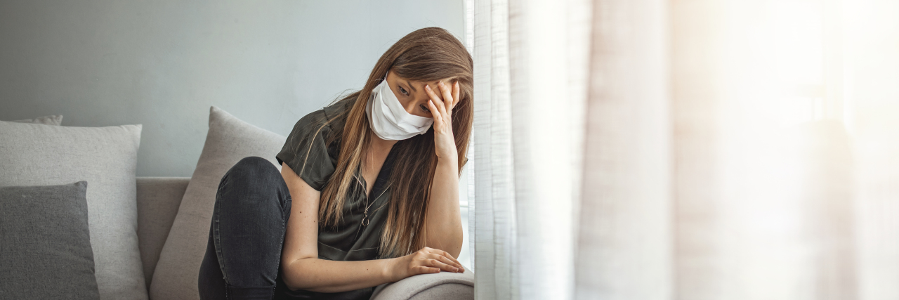 Managing Quarantine With Chronic Illness by Helping Others woman with long brown hair sitting on the couch with a mask on, hand on her head in exhaustion