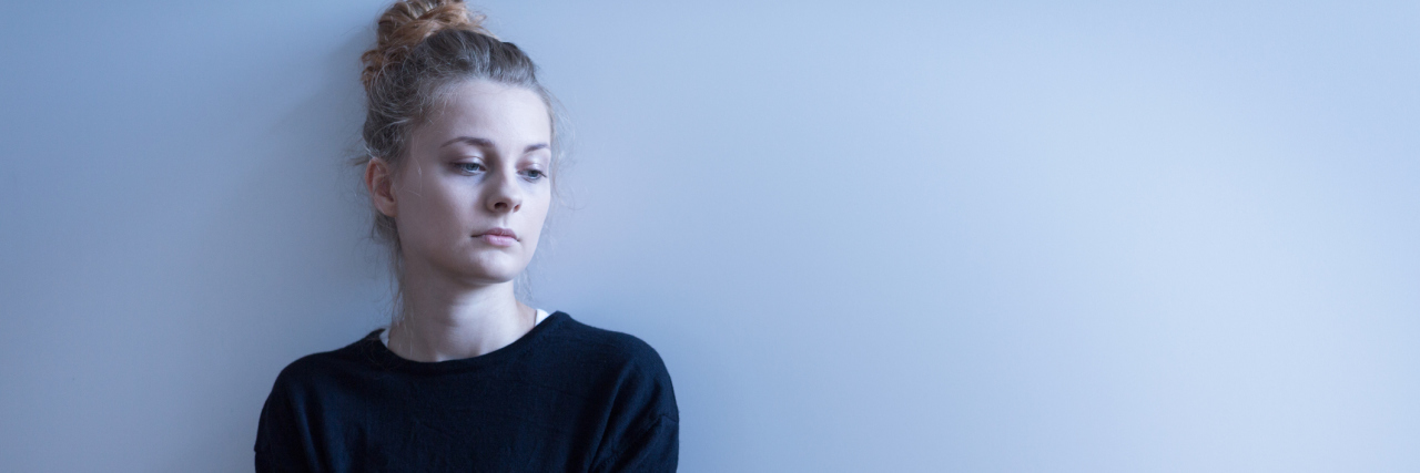 Living With Bipolar Disorder and Psychosis Feels Like Hell Young white woman sitting against the wall, sad with her arms folded
