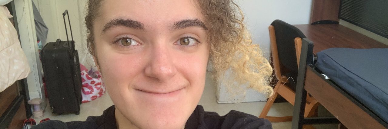My Transition to College During the Pandemic as a Disabled Teen A teenage girl with neat curly dark brown hair with a blond strand hanging taking a selfie highlighting her greenish-hazel eyes by sunlight.