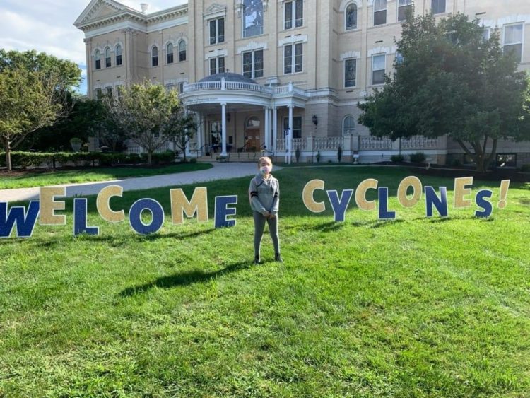 Standing outside on the grass in front of a school with a blue and gold sign that says "Welcome Cyclones."