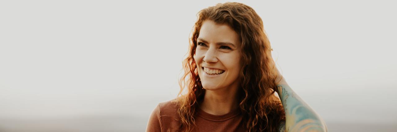 Learning It's OK to Show Both My Physical and Emotional Scars woman with curly red hair standing outside on a cloudy day with her tattooed arm scratching her head, smiling