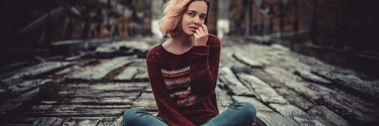 Asking 'Why Am I Like This?' With an Undiagnosed Mental Illness photo of woman sitting on a bridge looking into camera with one hand raised to her lips