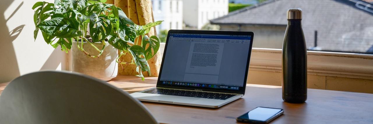 Work-From-Home Jobs for People With Disabilities and Chronic Illnesses An open laptop sits on a desk facing a window alongside a cellphone and water bottle