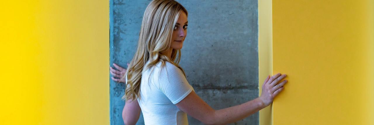 Can Your Period Cause Other Health Conditions? A young woman with long blonde hair wears jeans and a white T-shirt standing in the doorway of a yellow building