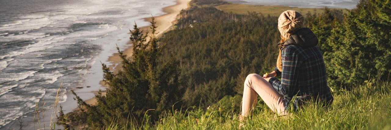 Photo of a white woman in a flannel shirt sitting on a hill overlooking water
