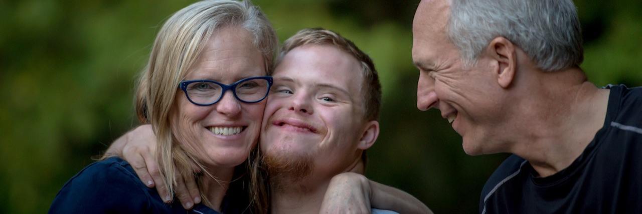 A young man with Down syndrome hugs his mother while his father stands nearby