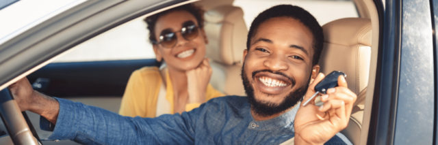 Young man holding a car key smiling out of a car window with a smiling woman in the background