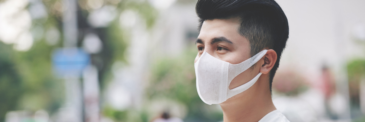 We Need to Talk About Chronic Illness and Suicide Young Asian man standing outside in a white long sleeve shirt with his arms folded, wearing a medical mask looking away