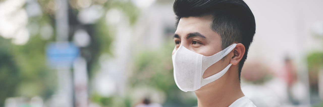 Young Asian man standing outside in a white long sleeve shirt with his arms folded, wearing a medical mask looking away