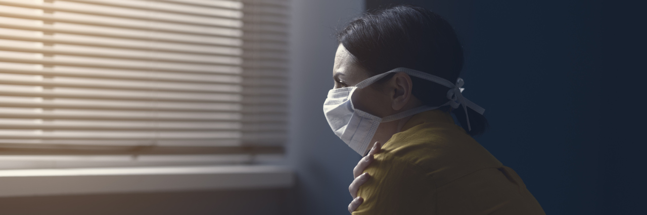 A middle-aged white woman wearing a surgical mask, sitting in a room alone, sad