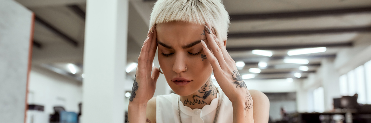 I'm a Disabled, Queer Abuse Survivor and I Can't Advocate Anymore young white woman with short blonde hair sitting at a desk with her elbows on the table, hands on the side of her head with eyes closed in frustration