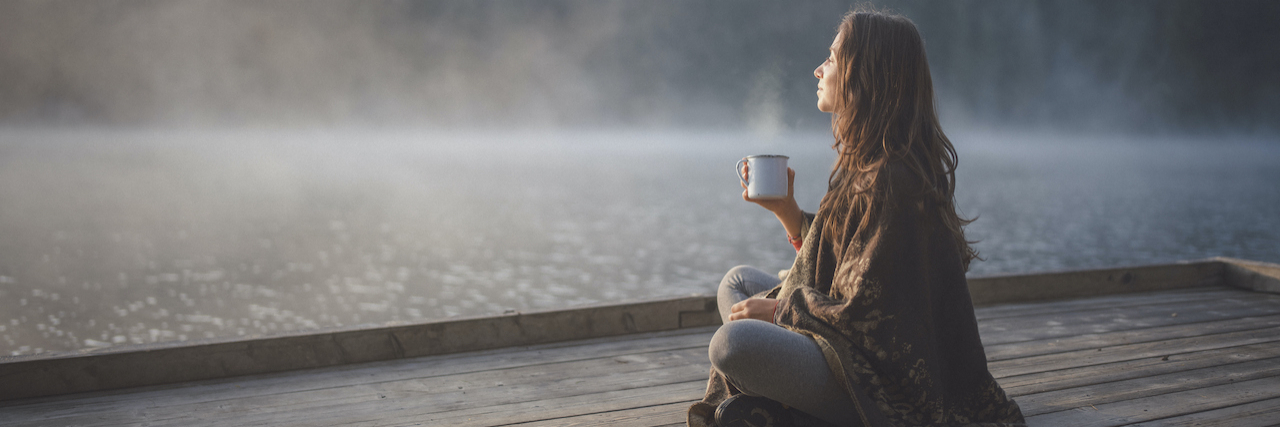 Woman sitting on wooden pier, drinking out of a mug and looking out at fog over the water