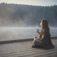 Woman sitting on wooden pier, drinking out of a mug and looking out at fog over the water