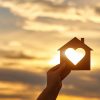 Hand holds wooden house in the form of heart against the sun.