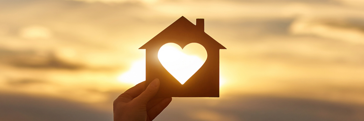 Sharing a Story of Homelessness and Chronic Illness Hand holds wooden house in the form of heart against the sun.