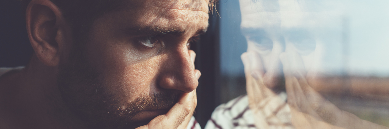 How Your Own Mind Might Be Gaslighting You Into Depression young white man with brown hair and a striped shirt leaning against a window with a sad expression, looking at his reflection