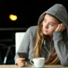sad young woman wearing a hooded sweatshirt in a cafe, with headphones and her phone, looking into distance