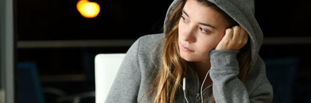 sad young woman wearing a hooded sweatshirt in a cafe, with headphones and her phone, looking into distance