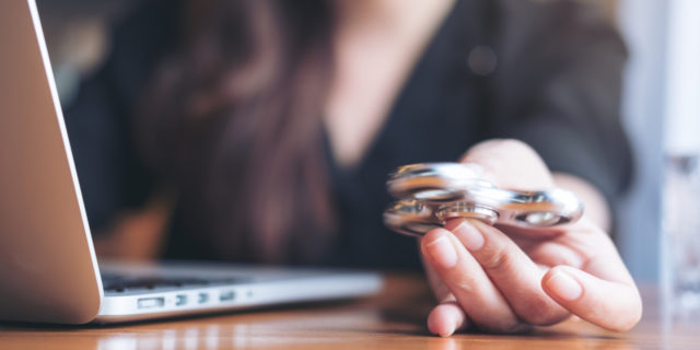 close up photo of woman playing with a fidget spinner at her desk beside her laptop