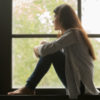 photo of young woman sitting by window looking out, knees raised
