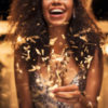 Cheerful Black woman holding single sparkler in hand outdoor.