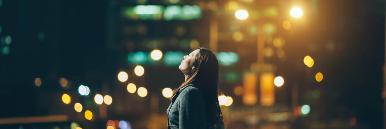 Woman standing in front of city buildings at night, looking up
