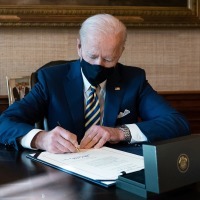 Joe Biden wears a mask while sitting behind a desk signing a bill into law