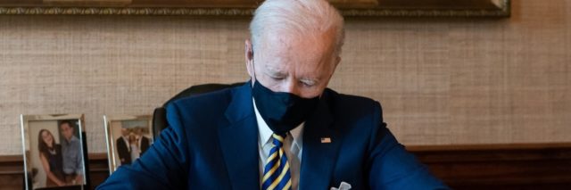 Joe Biden wears a mask while sitting behind a desk signing a bill into law
