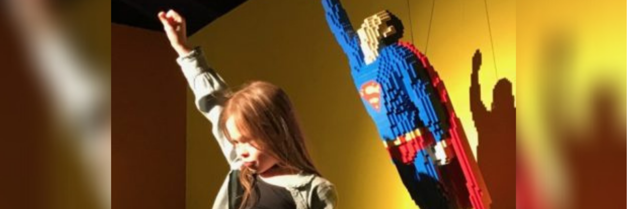 A photo of the author's daughter, a young white girl striking a superhero pose to match Superman in the background
