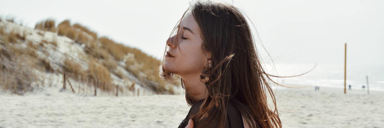 woman sitting on a sandy beach with hand on her arm, eyes closed with her head up and wind slightly blowing her hair