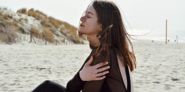 woman sitting on a sandy beach with hand on her arm, eyes closed with her head up and wind slightly blowing her hair