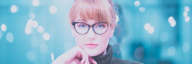 woman in glasses and a dark gray turtleneck looking at camera contemplatively with a hand on her chin