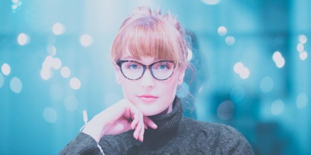woman in glasses and a dark gray turtleneck looking at camera contemplatively with a hand on her chin