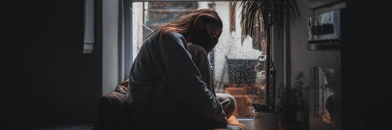 photo of woman sitting at home in front of a window, wearing a coronavirus mask and looking anxious
