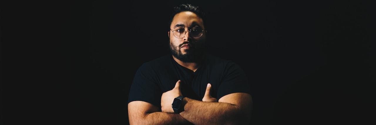 New Study Suggests Strong Link Between Autism and Substance Use Risk Man with dark hair, glasses and a black T-shirt stands with his arms folded against a black background