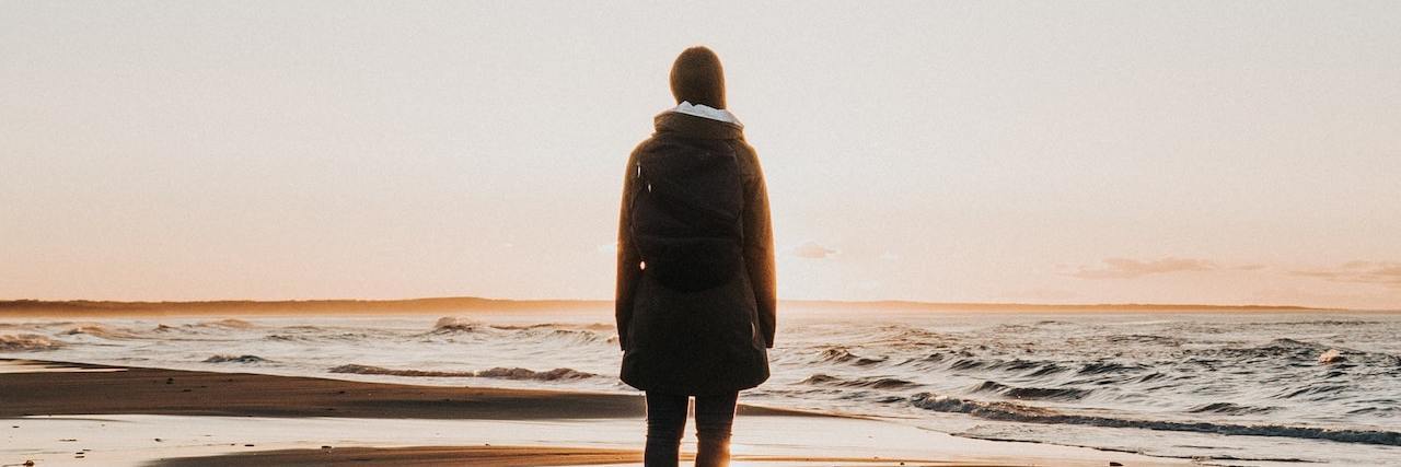 When Doctors Dismiss and Devalue Your Chronic Illness Woman standing on beach looking out, her reflection visible in the shallow water behind her
